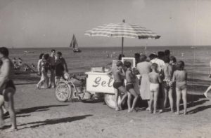Questa fotografia d'epoca in bianco e nero mostra una tipica e nostalgica scena estiva italiana del dopoguerra, con bagnanti in costume da bagno d'altri tempi in coda attorno a un carretto dei gelati motorizzato trainato da una motocicletta con sidecar/tre ruote direttamente sulla sabbia della spiaggia