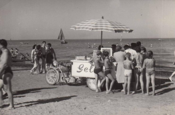 Questa fotografia d'epoca in bianco e nero mostra una tipica e nostalgica scena estiva italiana del dopoguerra, con bagnanti in costume da bagno d'altri tempi in coda attorno a un carretto dei gelati motorizzato trainato da una motocicletta con sidecar/tre ruote direttamente sulla sabbia della spiaggia